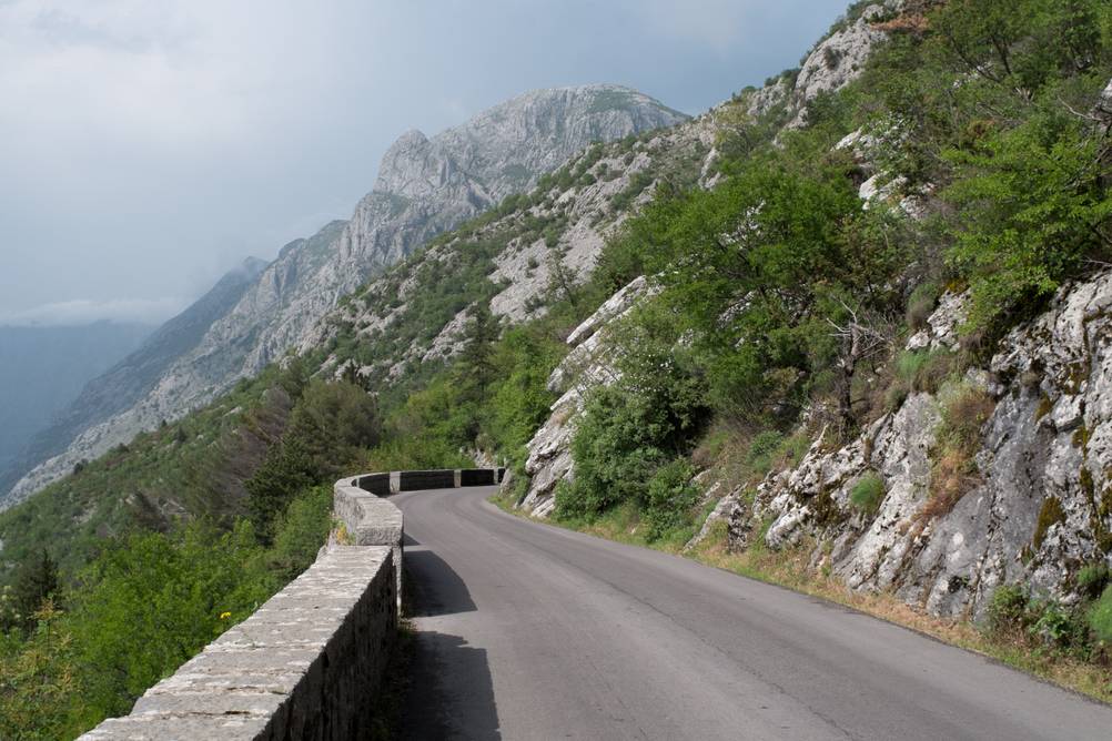 Ausschnitt der berühmten Straße P1 in Montenegro. Links trennt eine niedrige Steinmauer die Straße von einem Abhang, rechts entlang der Straße ist ein Berg. In der Perspektive ist ein Gebirge.