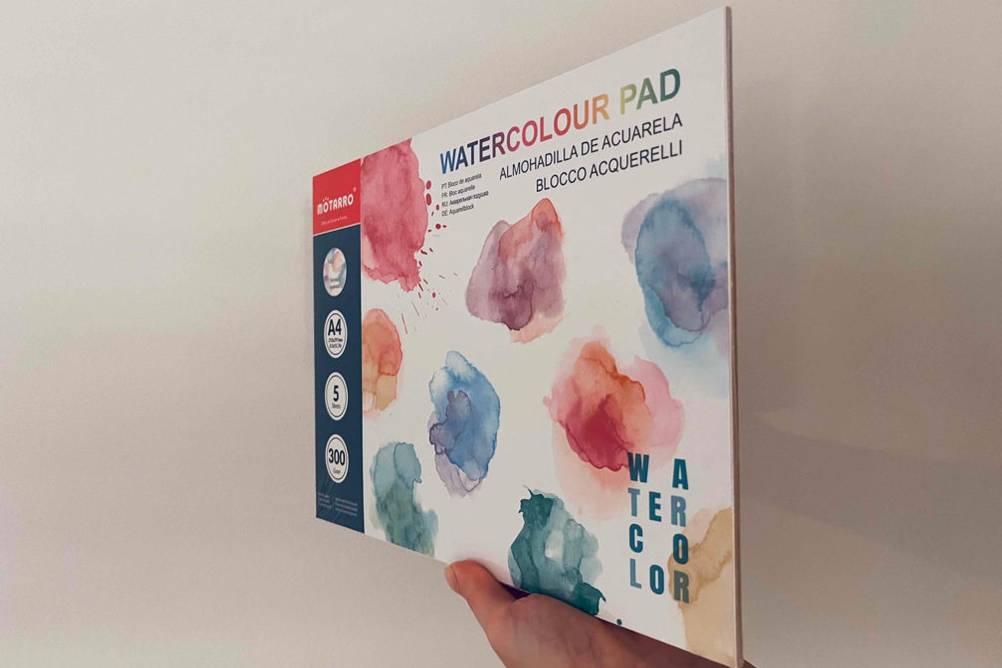 Hand hält getestetes Aquarellpapier von Motarro im Block mit bunten Aquarelltupfern hoch.