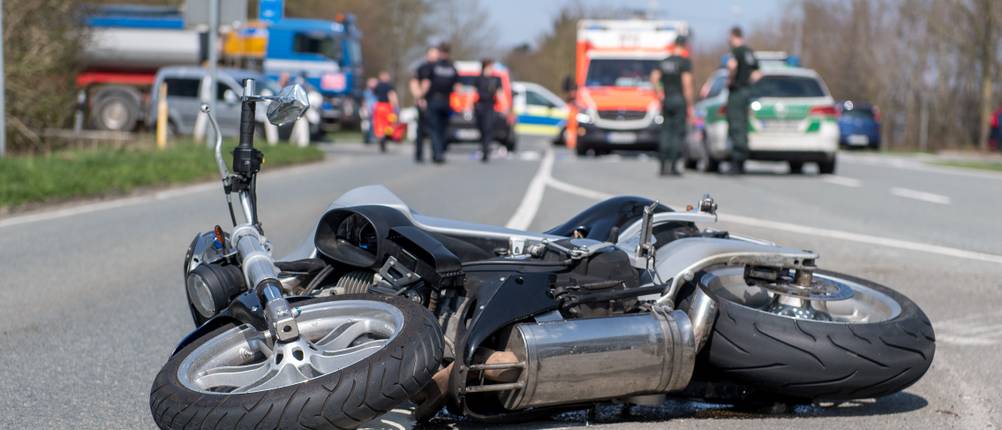 Motorradversicherung-Test: Ein silbernes Motorrad liegt auf der Straße, während im Hintergrund Polizisten und Sanitäter im Einsatz sind.
