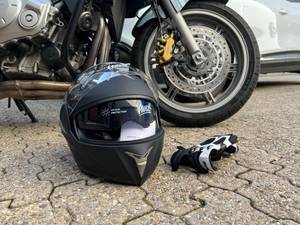 Motorradhelm liegt auf der Erde neben Motorradhandschuhen, vor einem Motorrad. 