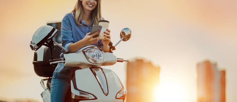 Eine Frau sitzt bei Sonnenuntergang auf einem Motorrad und hält einen Kaffeebecher und ein Smartphone in den Händen.