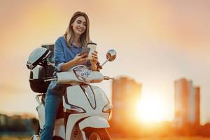 Motorradversicherung-Test: Eine Frau sitzt bei Sonnenuntergang auf einem Motorrad und hält einen Kaffeebecher und ein Smartphone in den Händen.