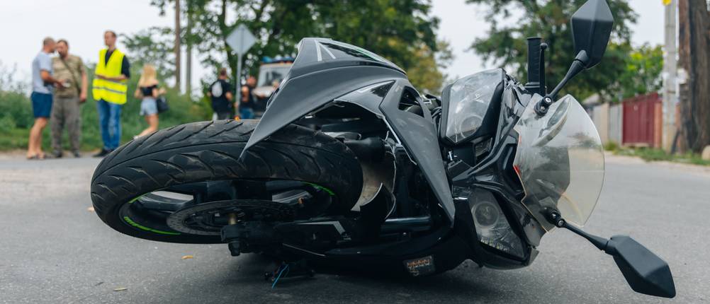 Motorradversicherung-Test: Ein schwarzes Motorrad liegt beschädigt auf der Straße, während im Hintergrund Menschen mit Warnwesten stehen.