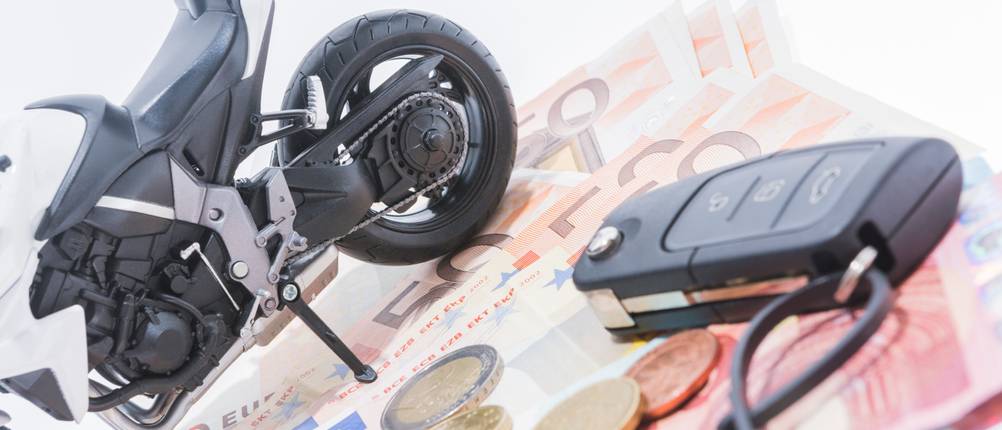 Auf einem Haufen von Geldscheinen steht ein Modell-Motorrad, während daneben ein Schlüssel liegt.