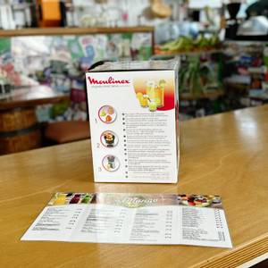 Seitenansicht der Moulinex PC603D10 Verpackung mit Produktinformationen und Funktionsbeschreibung. Das Bild zeigt das Gerät in einem Café-Umfeld auf einer Holztheke.