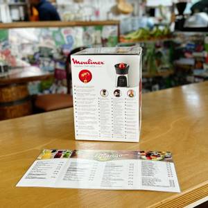 Die Moulinex PC603D10 Verpackung von hinten mit einer mehrsprachigen Beschreibung der Funktionen. Das Produkt steht auf einer Holztheke, im Hintergrund befindet sich eine Café-Einrichtung.