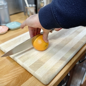 Eine Hand schneidet eine Orange auf einem Holzbrett mit einem großen Küchenmesser. Das Bild zeigt eine Vorbereitung für das Entsaften.