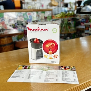 Die Verpackung der Moulinex PC603D10 Zitruspresse auf einer Holztheke mit einem Menü im Vordergrund. Im Hintergrund ist ein Café mit bunten Wänden und Regalen zu sehen.