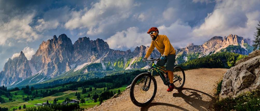 Mountainbike-Helm Herren im Test: Ein Mann trägt einen orange-roten Helm und fährt auf seinem Mountainbike auf einem Weg vor einer Bergkulisse.