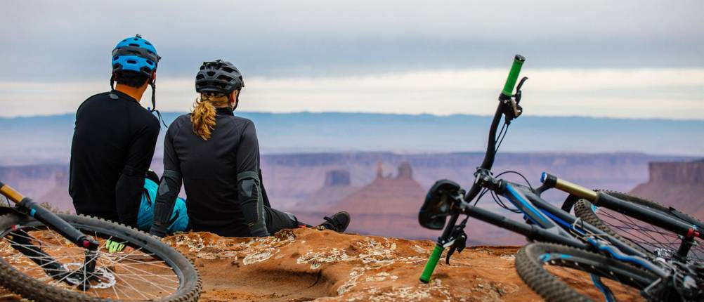 Mountainbike-Helm Herren Test: Ein Mann und eine Frau schauen sitzend auf eine Canyon-Landschaft. Beide tragen einen Helm. Ihre Mountainbikes haben sie auf den Boden gelegt.