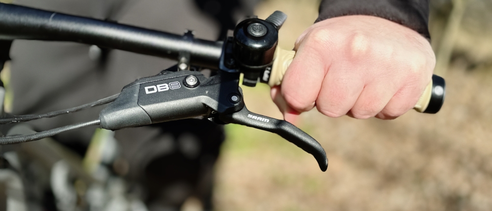 Eine Hand umfasst einen Fifty-Fifty-MTB-Griff während der Fahrt. Der schwarze Lenker mit SRAM DB8 Bremshebel ist im Fokus, der Hintergrund zeigt einen Trail.
