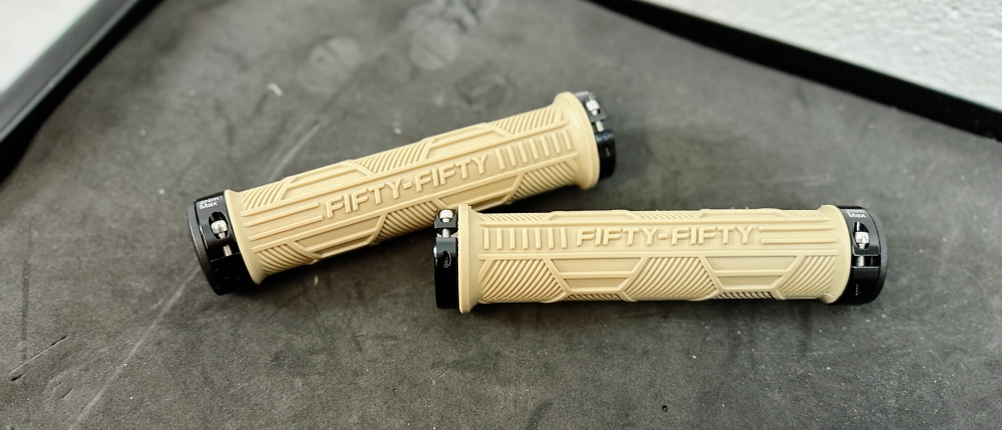Zwei beige FIFTY-FIFTY MTB-Griffe liegen auf einer dunklen Oberfläche.