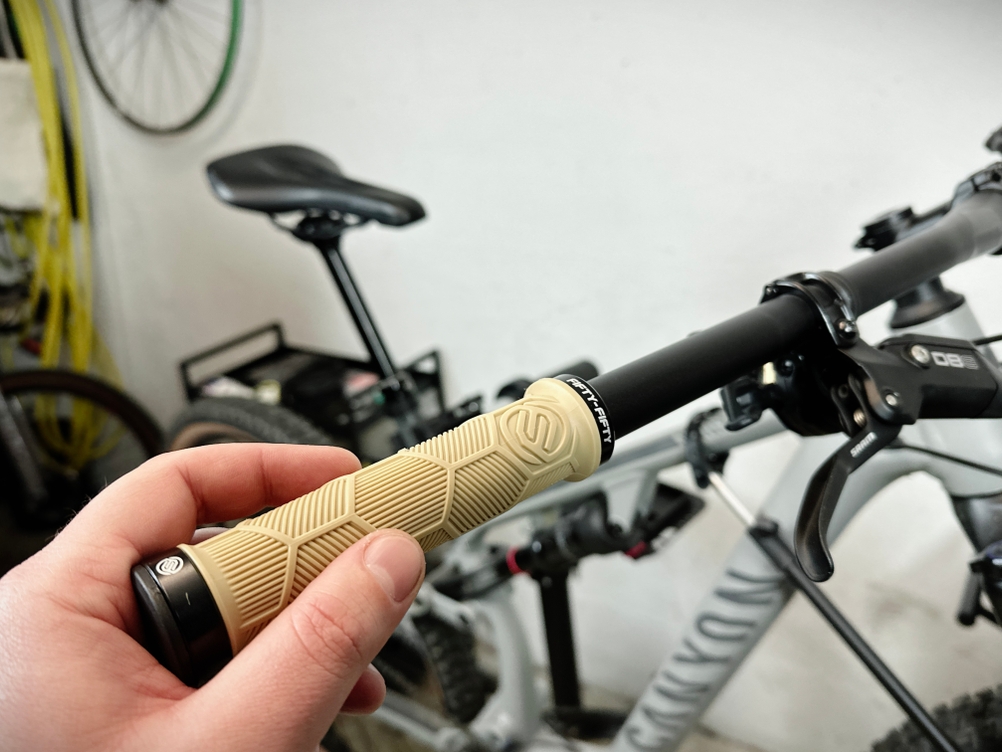 Ein beiger FIFTY-FIFTY MTB-Griff wird an einem schwarzen Lenker montiert. Eine Hand hält den Griff fest, während das Fahrrad im Hintergrund steht.