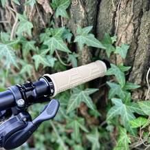 Ein beiger MTB-Griff der Marke FIFTY-FIFTY, montiert an einem schwarzen Lenker, lehnt an einem Baum mit Efeu. Der Hintergrund zeigt Naturdetails.