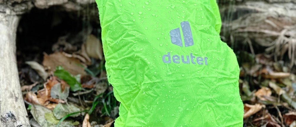Der MTB-Rucksack von Deuter ist mit Schutzhülle versehen, lehnt an einem Baumstamm im Wald und wird beregnet.