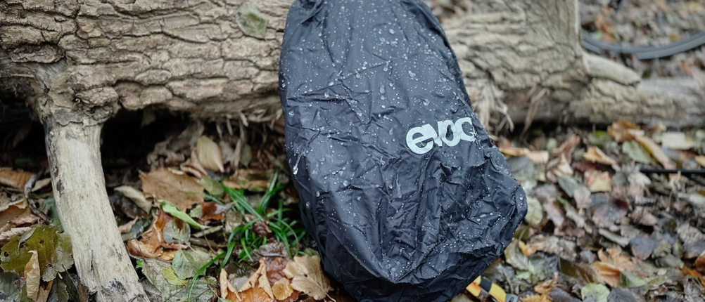 Der MTB-Rucksack von Evoc ist mit Schutzhülle versehen, lehnt an einem Baumstamm im Wald und wird beregnet.