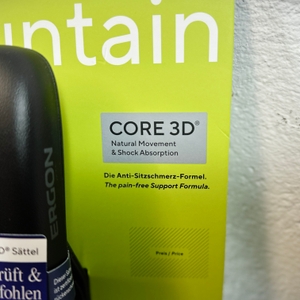 Der Ergon SMC Core MTB-Sattel vor einer gelben Verpackung mit dem Hinweis auf die Core 3D-Technologie und die Anti-Schmerz-Formel. Ein Teil des Sattels ist im Vordergrund zu sehen.