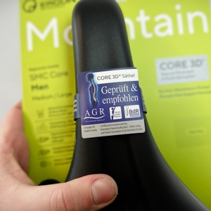 Ein Ergon SMC Core MTB-Sattel wird in der Hand gehalten. Ein AGR-Siegel mit dem Hinweis „Geprüft & empfohlen“ ist auf dem Sattel angebracht.