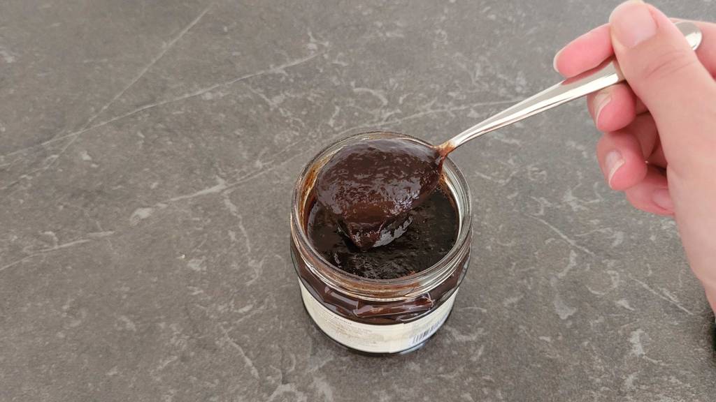Hand hält einen Teelöffel mit getestetem Pflaumenmus über ein Glas mit Pflaumenkompott.