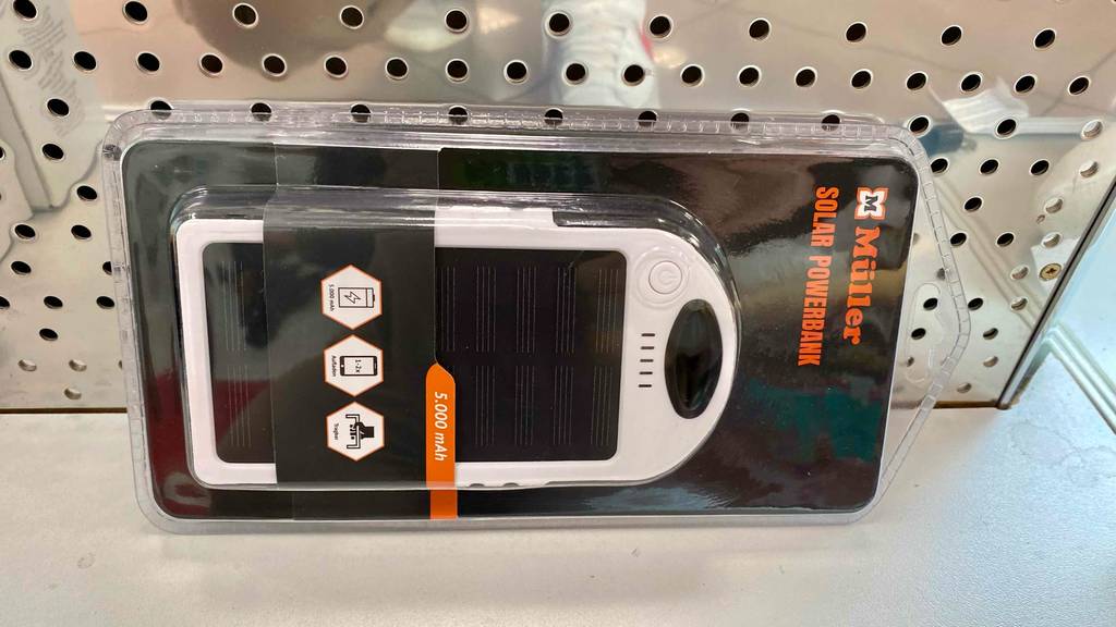 Eine transparente Müller Kunststoffverpackung mit einer Solar-Powerbank im Test in Weiß liegt vor einer gelöcherten Metallfläche.