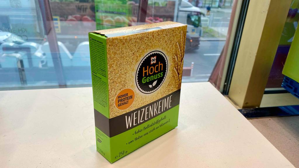 Ansicht von der Seite inklusive Produktinformationen einer gelb-schwarz-grünen Verpackung mit Müller Hoch Genuss Weizenkeime im Test vor einem Schaufenster mit Blick nach draußen.