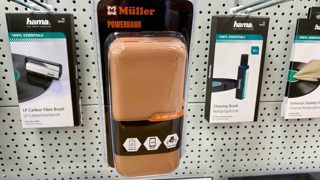 An einem Verkaufsregal hängt eine lachsfarbene Powerbank mit 20.000 mAh im Test, die sich in einer transparenten Verpackung von Müller neben weiterem Elektronik-Zubehör befindet.
