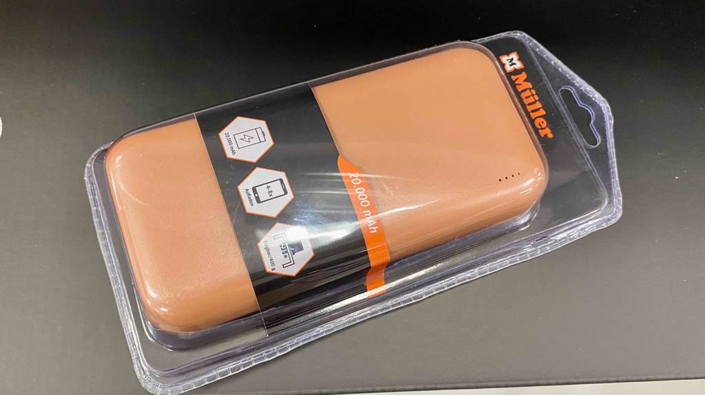 Eine lachsfarbene Powerbank mit 20.000 mAh im Test liegt in ihrer transparenten Verpackung mit dem Markenzeichen Müller auf einer dunkelgrauen Fläche.