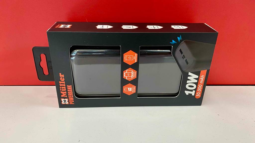 Eine schwarz-orange Müller Verpackung inklusive einer schwarzen Powerbank mit 30.000 mAh im Test liegt auf der Seite auf weißer Fläche vor einer roten Wand.