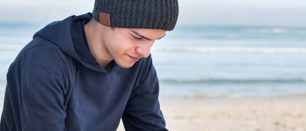 Mütze-mit-Licht-Test: Mann sitzt am Strand und trägt dunkelblauen Hoodie mit grauer Mütze mit Bluetooth.