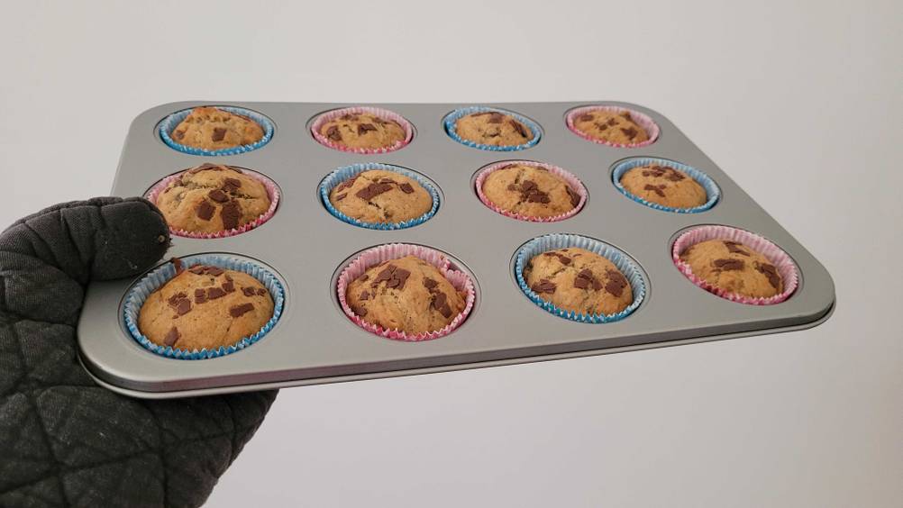 Eine Hand hält eine getestete Muffinform mit gebacken Muffins vor eine helle Wand.