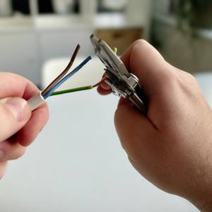 Zwei Hände nutzen das Leatherman Skeletool, um ein Elektrokabel abzuisolieren. Drei freigelegte Adern in Braun, Blau und Gelb-Grün sind sichtbar.