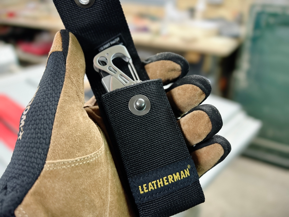 Eine Hand mit Arbeitshandschuh hält das Leatherman Skeletool in einer schwarzen Gürteltasche. Der Hintergrund zeigt eine Werkstatt mit verschiedenen Werkzeugen.