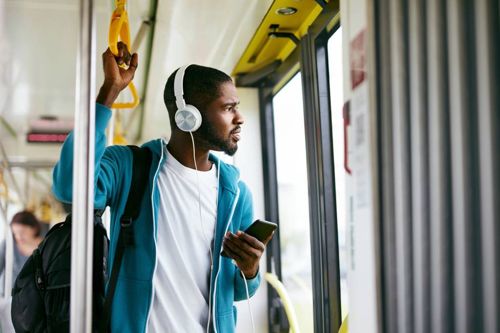 Ein junger Mann in einer blauen Jacke steht im Zug und schaut aus dem Fenster, während er Musik hört.