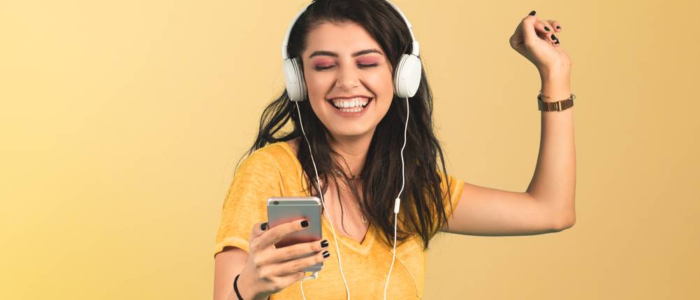 Musik-Streaming-Test: Eine junge Frau steht vor einem gelben Hintergrund und tanzt, während Sie Kopfhörer trägt.