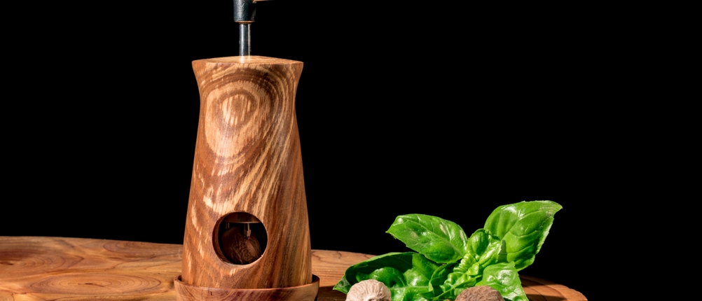 Muskatmühlen-Test: Muskatmühle aus Holz steht auf einem Holztisch vor einem schwarzen Hintergrund. Daneben liegen zwei Muskatnüsse und Basilikum.