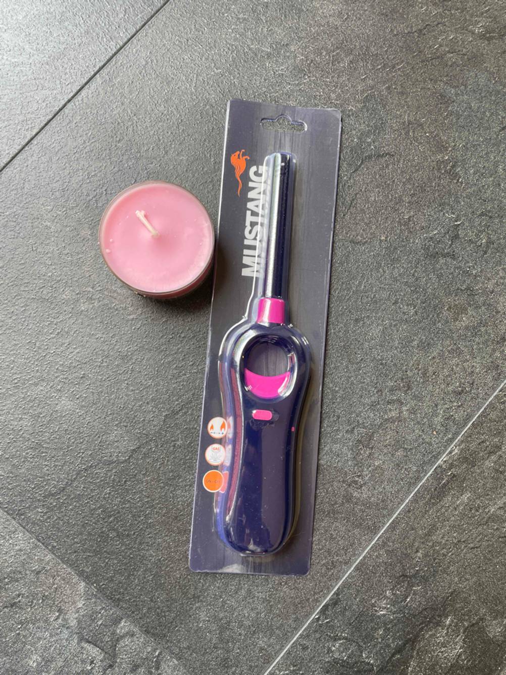 Stabfeuerzeug im Test: ein Feuerzeug in Originalverpackung liegt neben einem rosa Teelicht auf schwarzen Fliesen.