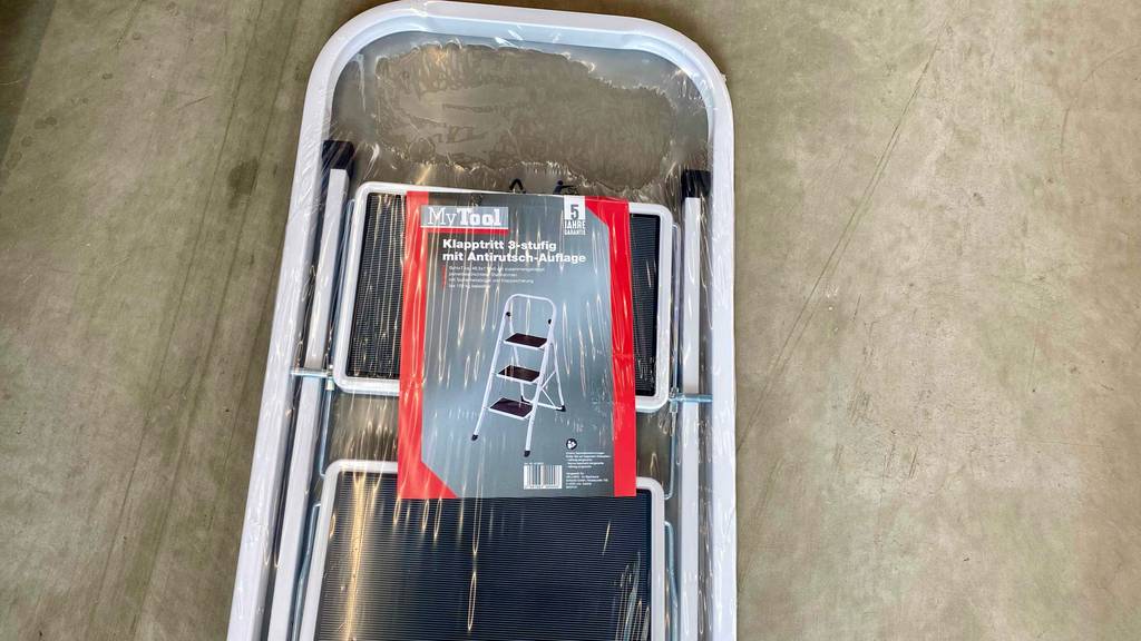 Detailansicht des oberen Bereichs einer weißen 3-Stufen-Trittleiter im Test mit schwarzen, gummierten Trittflächen, die innerhalb der transparenten Kunststoffverpackung an einer grauen Wand lehnt.