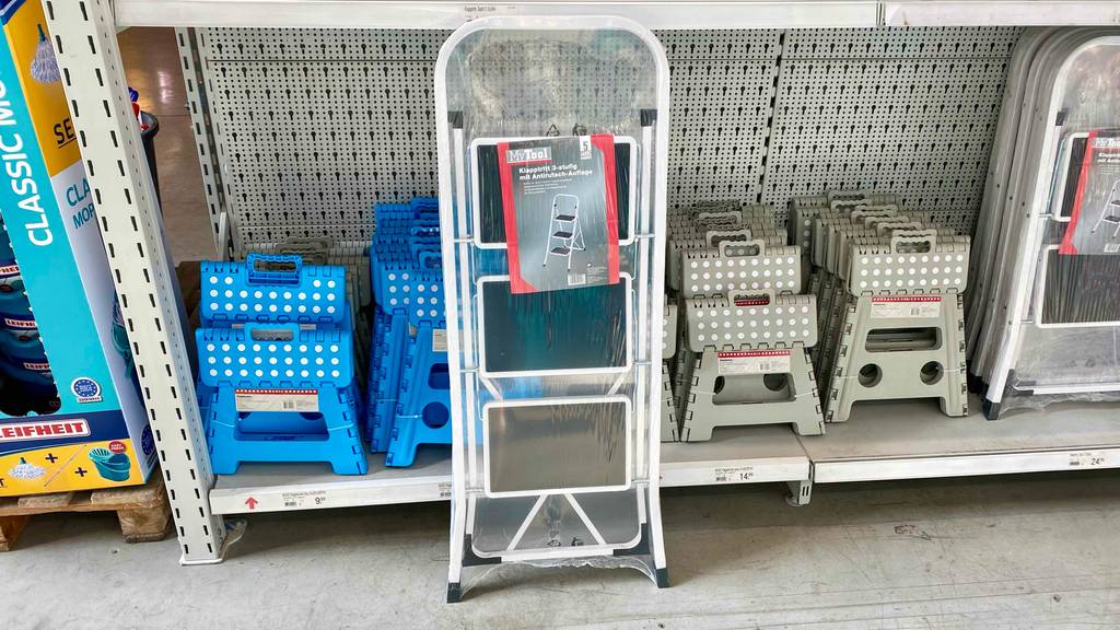 Eine getestete Trittleiter mit 3 Stufen steht vor einem Regal mit verschiedenen Leitern in einem Baumarkt.