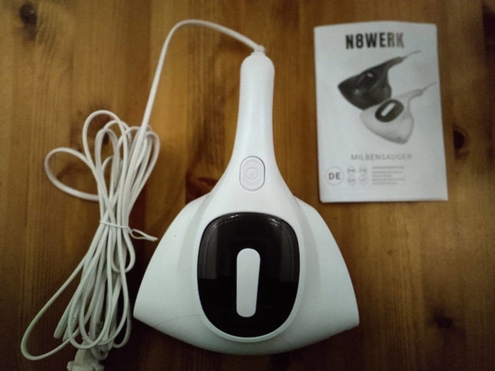 Weißer N8Werk Milbenstaubsauger im Test liegt neben der Bedienungsanleitung auf einem Holzuntergrund.