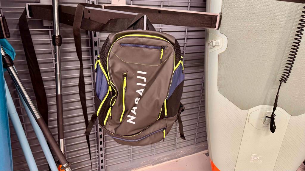 Ein dunkler Schwimmrucksack im Test hängt an einem Verkaufsregal zwischen Walking-Stöcken und einem aufblasbaren Stand-up-Paddling Board.