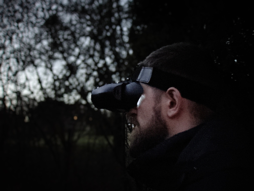 Ein Mann trägt das Bresser-Digital-NV-Binokular 1x mit Kopfhalterung im Wald bei Dämmerung. Das Nachtsichtgerät ist fest über den Augen positioniert.