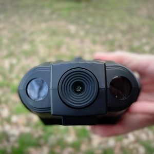 Vorderansicht des Bresser-Digital-NV-Binokulars mit zwei Linsen und einem zentralen Infrarotsensor. Eine Hand hält das Gerät, der Hintergrund zeigt eine unscharfe Wiese.