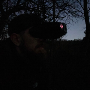 Ein Mann trägt das Nachtsichtgerät Nightfox Swift mit Kopfhalterung im Wald bei Dämmerung. Die rote IR-Leuchte des Geräts ist gut sichtbar.