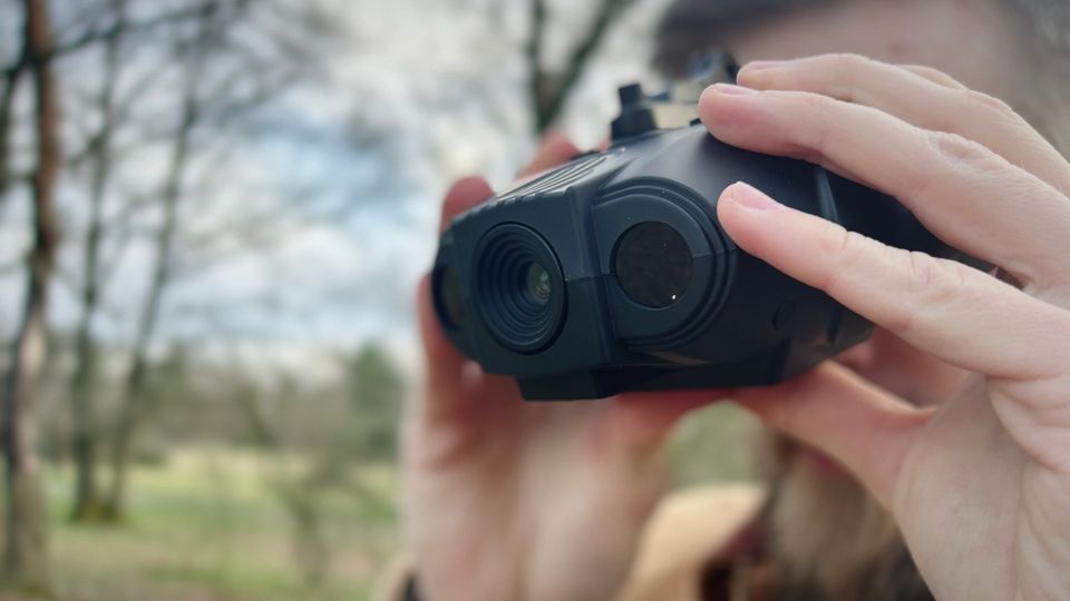 Ein Mann nutzt das Nightfox Swift Nachtsichtgerät im Freien. Er hält es mit beiden Händen vor die Augen, im Hintergrund sind Bäume und eine Wiese.