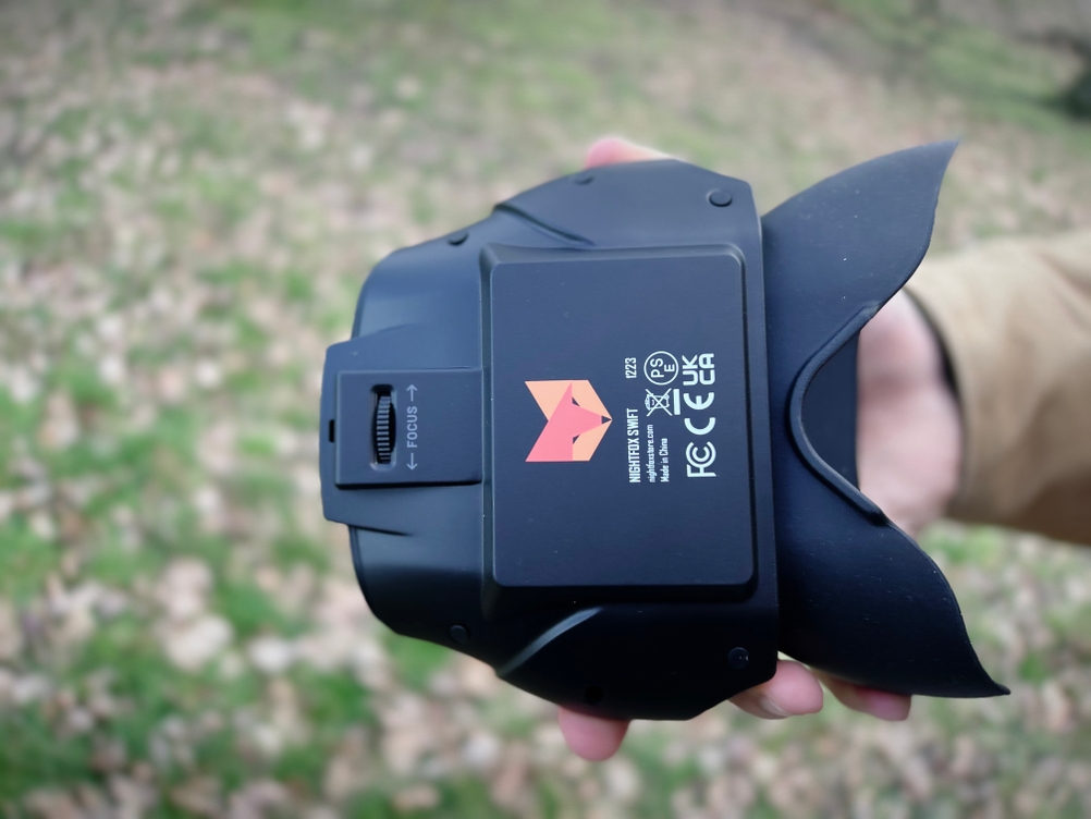 Unterseite des Nightfox-Swift-Nachtsichtgeräts mit Fokusrad und Nightfox-Logo. Eine Hand hält das Gerät, der Hintergrund ist unscharf.