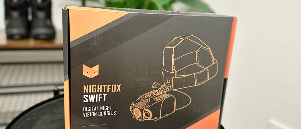 Die Verpackung des Nightfox-Swift-Nachtsichtgeräts steht auf einem Tisch, im Hintergrund sind Pflanzen und Schuhe. Das Design zeigt das Gerät mit Kopfhalterung und das Nightfox-Logo.