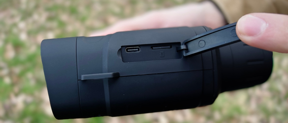 Eine geöffnete Abdeckung zeigt die USB- und TF-Karten-Anschlüsse des Relassy-NV4000-Nachtsichtgeräts. Eine Hand hält das Gerät, der Hintergrund ist unscharf.