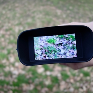 Das Relassy NV4000 Nachtsichtgerät mit eingeschaltetem Display zeigt eine vergrößerte Ansicht des Bodens. Eine Hand hält das Gerät, der Hintergrund ist unscharf.