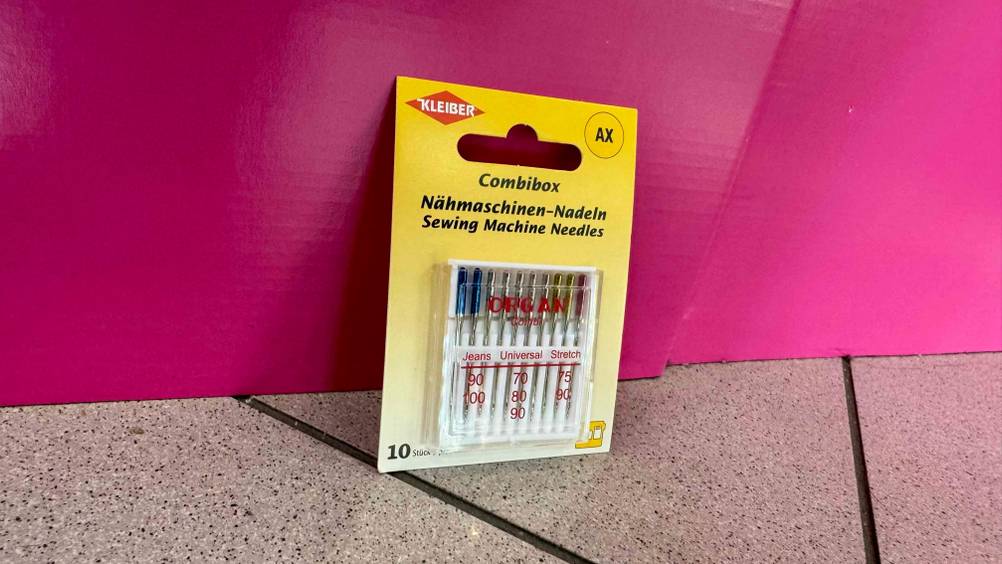 Eine Packung der getesteten Nähmaschinennadeln vor einer pinken Fläche auf dem Boden.