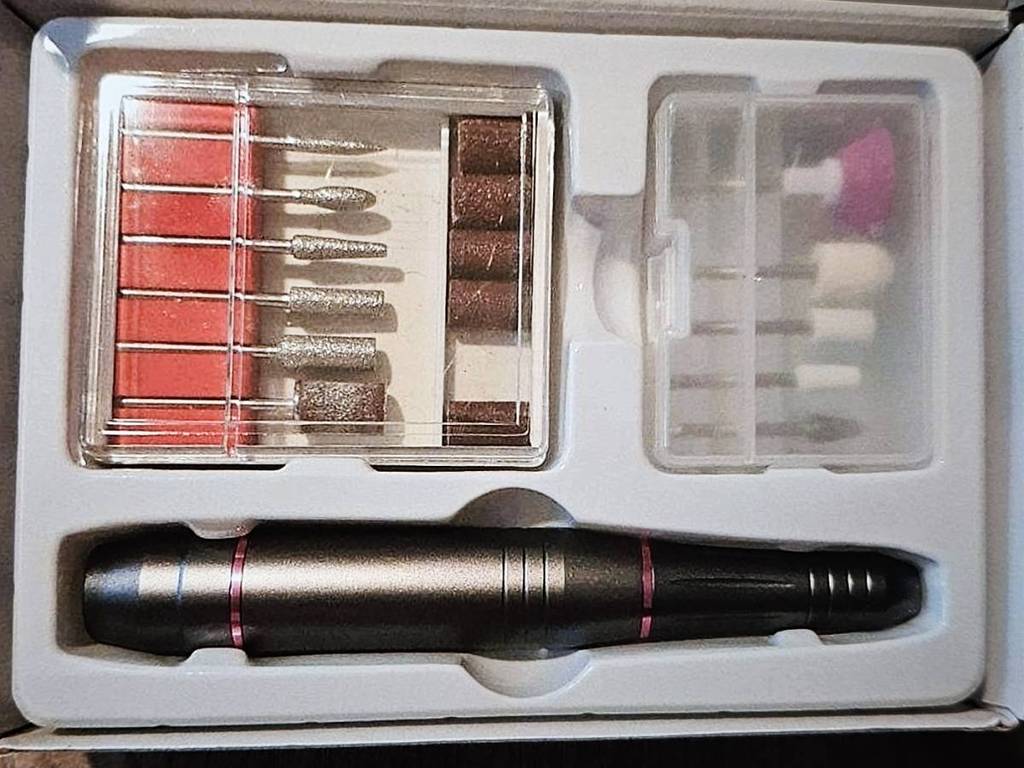 Nahaufnahme einer offenen Verpackung eines Maniküren-Sets inklusive eines elektrischen, schwarzen Maniküre-Stifts sowie verschiedenen Maniküre-Aufsätzen in zwei transparenten Kunststoff-Aufbewahrungsdosen.